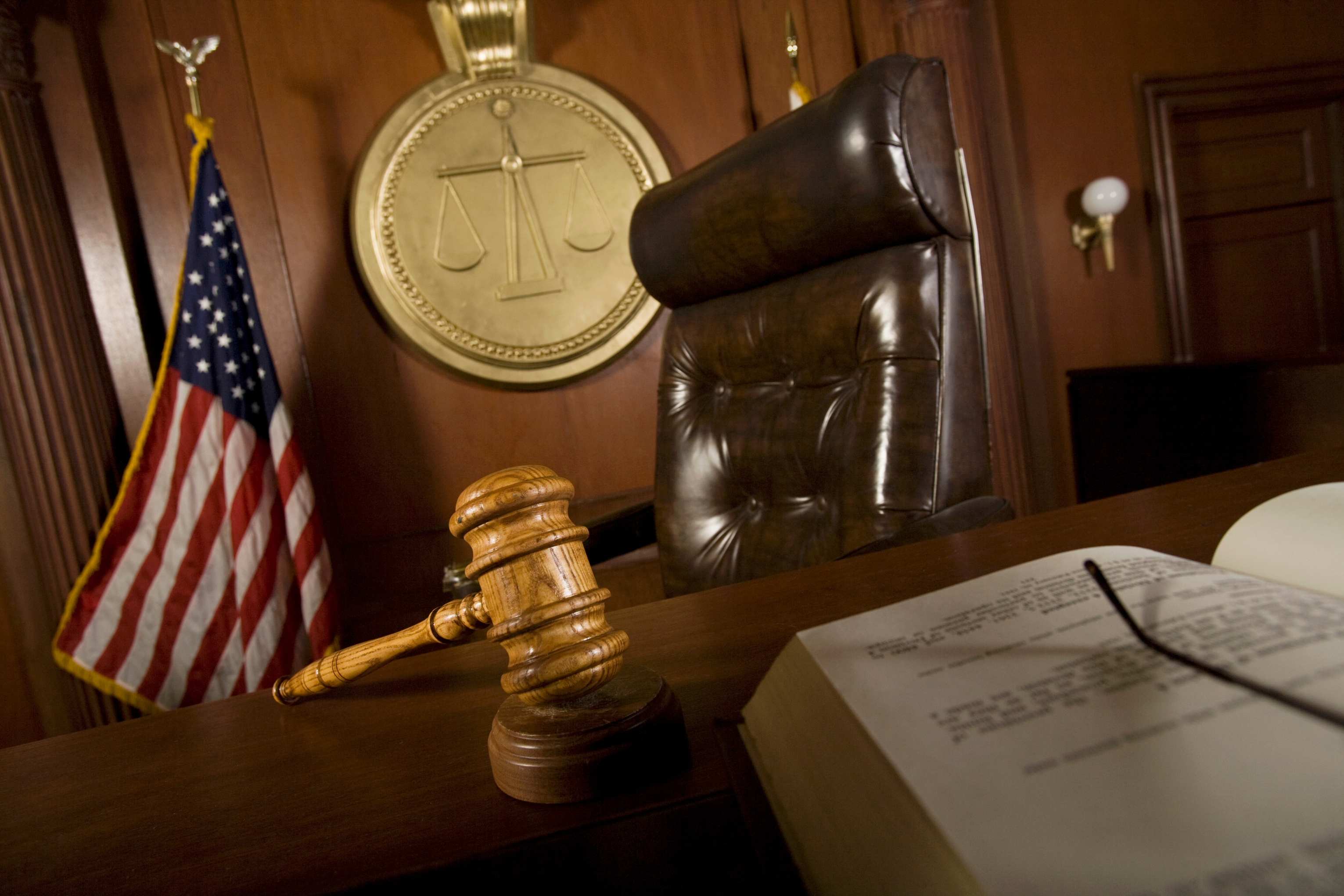 A judge's bench with a wooden gavel, American flag, and scales of justice emblem in a courtroom